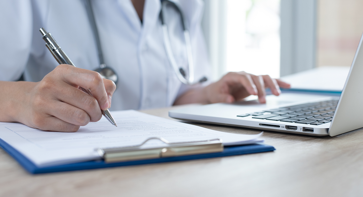 Healthcare professional in white coat with stethoscope writing on documents while using laptop computer at desk