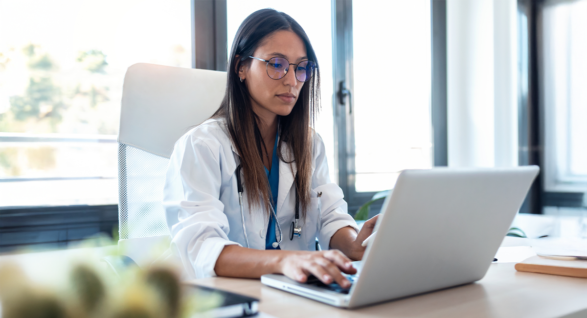 Female physician in white coat working on laptop at desk in modern office