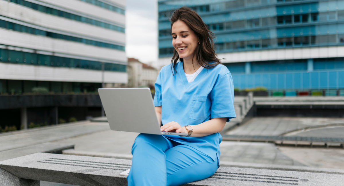 A smiling healthcare worker in scrubs sits outdoors on a bench, using a laptop