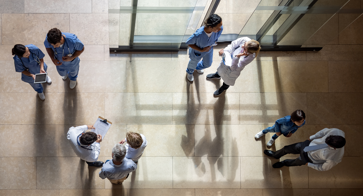 Overhead view of healthcare professionals in scrubs and white coats having informal discussions in small groups in a hospital hallway with beige tile flooring and natural light streaming through windows.