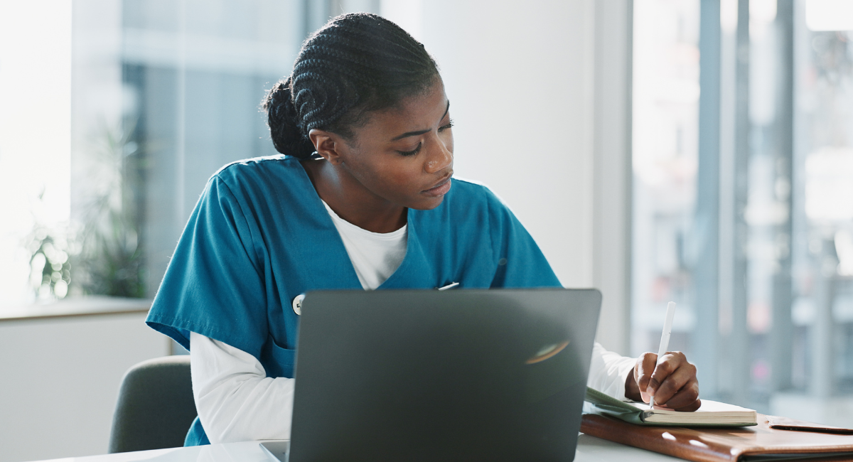 Nurse in blue scrubs working on laptop and taking notes at desk